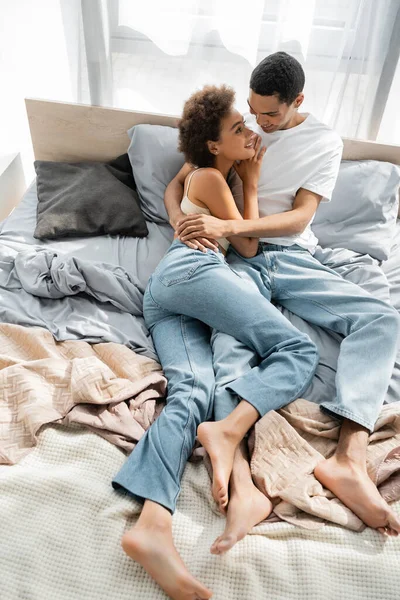 Blick aus der Vogelperspektive auf barfüßiges und glückliches afrikanisch-amerikanisches Paar in Jeans, das sich zu Hause auf dem Bett umarmt — Stockfoto