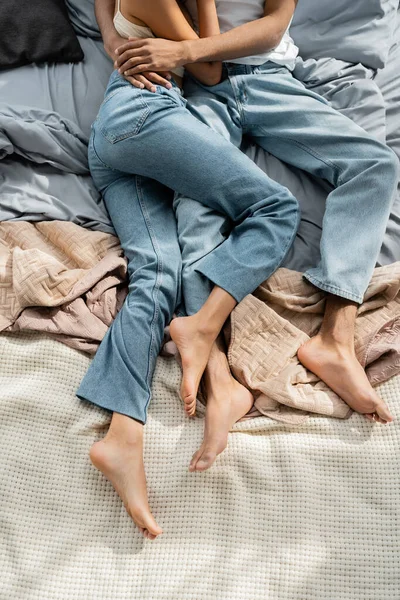 Сверху вид обрезанной босиком африканской пары в джинсах лежащей и обнимающейся на кровати — стоковое фото