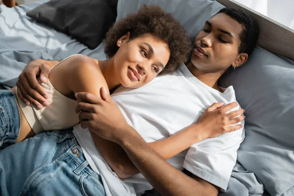 Femme afro-américaine souriante étreignant petit ami en t-shirt blanc sur le lit à la maison — Photo de stock
