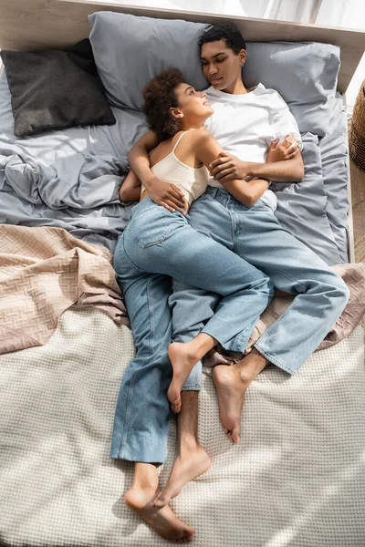 Зверху ви бачите африканську пару босоногих в джинсах, які обіймаються і дивляться один на одного на ліжку. — стокове фото