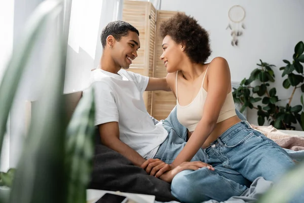 Sexy Africano americano casal sentado na cama e sorrindo uns para os outros em primeiro plano borrado — Fotografia de Stock