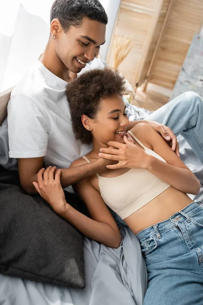 Улыбающийся африканский американец обнимает сексуальную девушку в топе и джинсах на кровати — стоковое фото