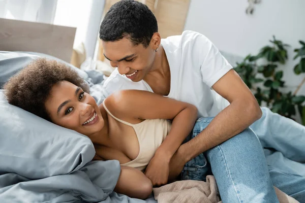 Cheerful african american woman looking at camera near smiling man hugging her on bed — Stock Photo
