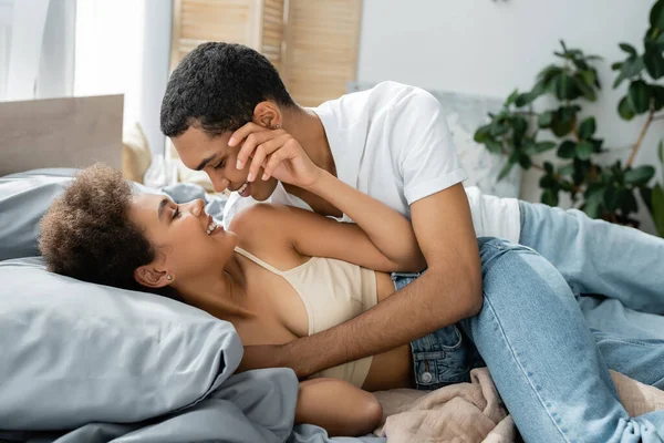 Sonriente afroamericano hombre abrazando apasionada novia tocando su cara en la cama - foto de stock