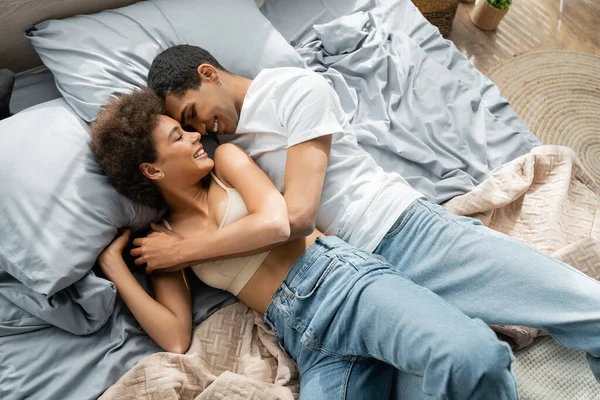 Vista superior de pareja afroamericana en jeans acostados en la cama y sonriendo con los ojos cerrados - foto de stock
