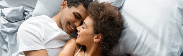 Top view of young african american couple lying on bed and smiling face to face with closed eyes, banner — Stock Photo