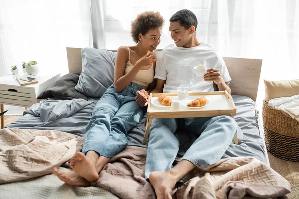 Young african american couple in jeans drinking coffee near tray with croissants on bed — Stock Photo