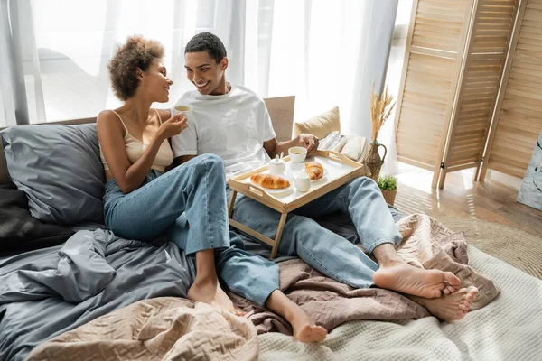 Полная длина босиком африканской пары в джинсах сидя рядом с подносом с кофе и круассанами на кровати — стоковое фото