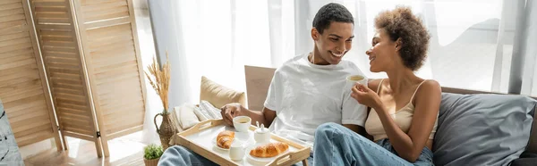 Jovem e feliz casal afro-americano sorrindo perto do café da manhã com café e croissants na cama, banner — Fotografia de Stock