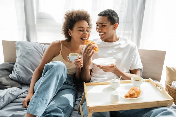 African american man in white t-shirt feeding girlfriend with croissant while having breakfast on bed — Stock Photo