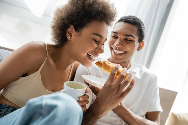 Joyful african american man feeding smiling girlfriend with croissant during breakfast in bedroom — Stock Photo