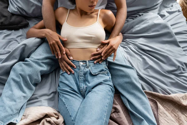 Ansicht von oben der beschnittenen afrikanisch-amerikanischen Frau in Jeans und bauchfreiem Top in der Nähe ihres Freundes, der sie auf dem Bett umarmt — Stockfoto