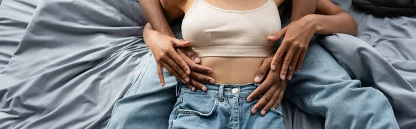 Partial view of african american man hugging sexy woman in crop top and jeans on bed, banner — Stock Photo