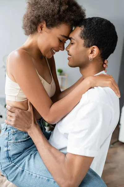 Seitenansicht eines afrikanisch-amerikanischen Mannes im weißen T-Shirt und einer sexy Frau im bauchfreien Top, die sich zu Hause mit geschlossenen Augen umarmt — Stockfoto