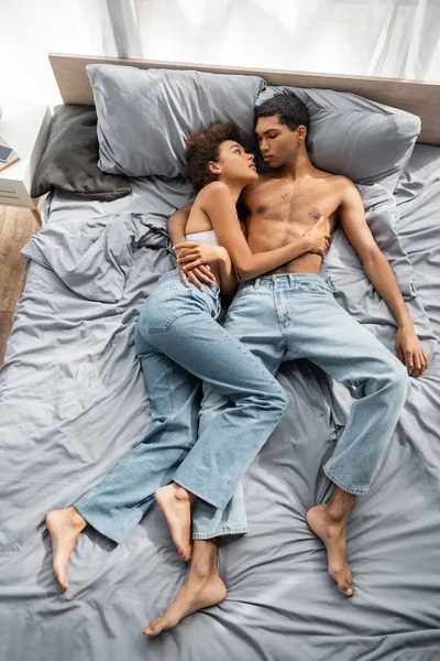 Hochwinkelaufnahme eines barfüßigen afrikanisch-amerikanischen Paares in Jeans, das auf dem Bett liegt und einander ansieht — Stockfoto