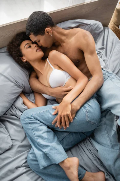 Top view of african american woman in bra and shirtless man lying on bed in jeans and looking at each other — Stock Photo