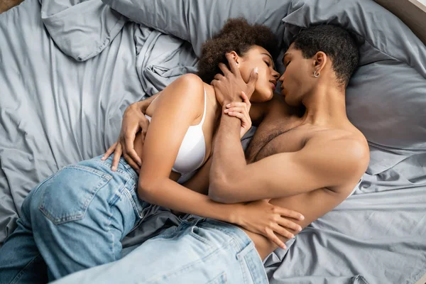 Вид сверху на африканского мужчину без рубашки, обнимающего горячую женщину в джинсах и лифчике на кровати — стоковое фото