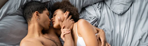 Top view of african american man touching face and embracing sexy girlfriend on bed, banner — Stock Photo
