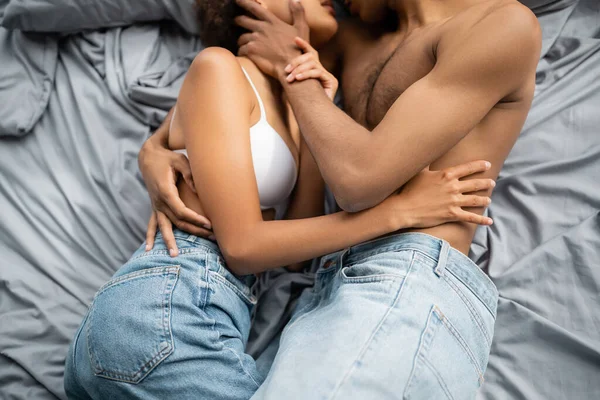 Обрезанный вид на мускулистого африканского мужчину, обнимающегося с девушкой в джинсах и лифчике на серой кровати — стоковое фото