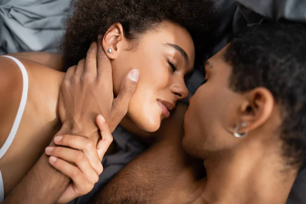 Vista superior del hombre afroamericano tocando la cara de la novia bonita y sexy en el dormitorio - foto de stock