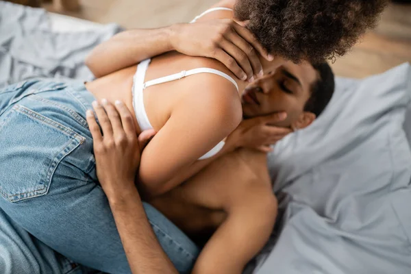 Hombre afroamericano borroso abrazando a mujer seductora en sujetador y jeans en la cama en casa - foto de stock