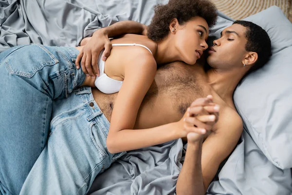 Пристрасна афроамериканська пара в джинсах, що тримаються за руки і дивляться один на одного на ліжку. — стокове фото