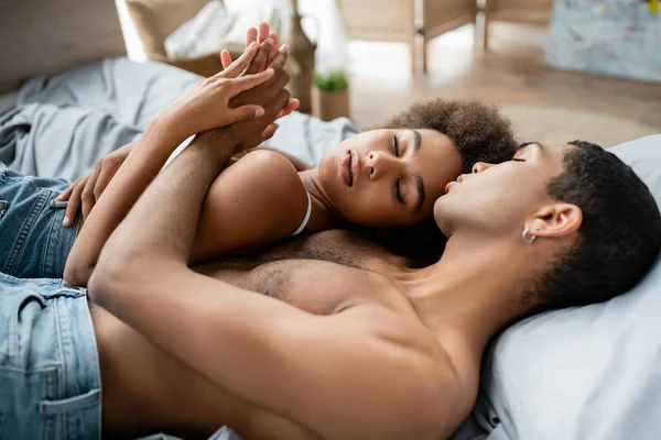 Joven y tierna pareja afroamericana acostada en la cama con los ojos cerrados y cogidas de la mano - foto de stock