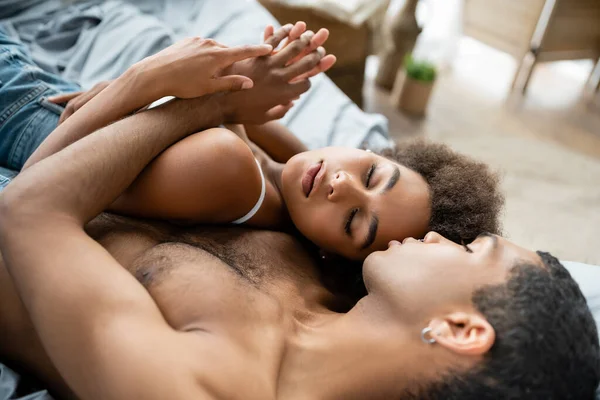 Mujer americana africana bonita con los ojos cerrados cogidos de la mano con el novio sin camisa en la cama - foto de stock