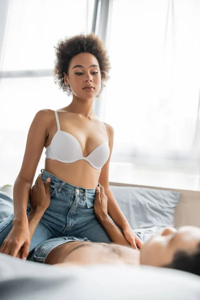 Hot african american woman in jeans and white bra near blurred man hugging her hips — Stock Photo