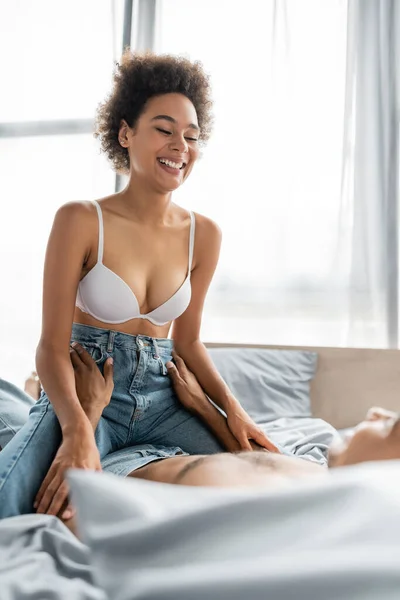 Felice e sexy donna africana americana in jeans e reggiseno sopra l'uomo sfocato sdraiato sul letto — Foto stock
