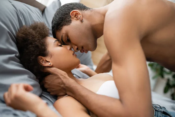 Vista lateral de shirtless muscular homem beijando afro-americano namorada na cama em casa — Fotografia de Stock