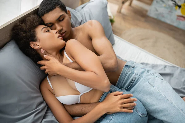 Вид зверху без сорочки афроамериканський чоловік і жінка в білому бюстгальтері, що ховається на ліжку, лежачи в джинсах — стокове фото