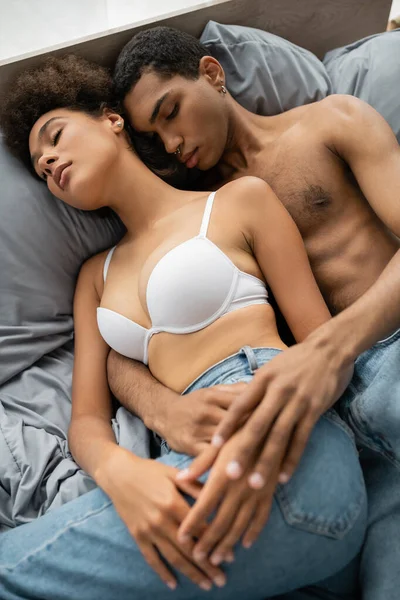 Высокий угол зрения на безрубашечного африканского мужчину и женщину в белом лифчике лежащих на кровати с закрытыми глазами — стоковое фото