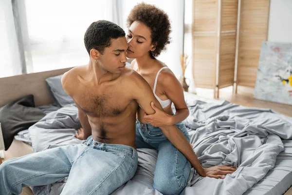 Африканский мужчина без рубашки сидит на кровати в джинсах рядом с сексуальной девушкой обнимая его на кровати — стоковое фото