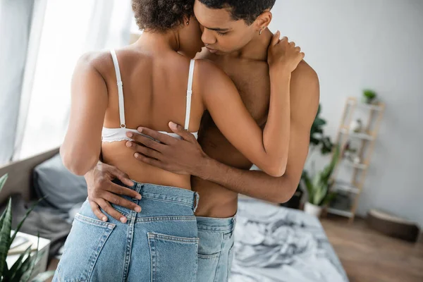 Joven y sin camisa afroamericano hombre abrazando novia seductora en sujetador y jeans - foto de stock