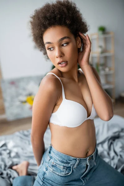 Sensual african american woman in white bra and jeans fixing curly hair while posing at home — Stock Photo