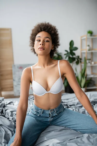 Bonita mujer afroamericana con cuerpo sexy posando en sujetador blanco y jeans en el dormitorio - foto de stock