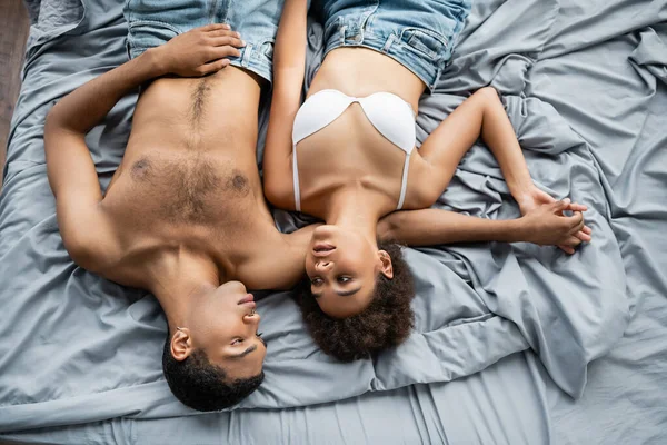 Top view of sexy african american lovers holding hands and looking at each other on grey bedding — Stock Photo