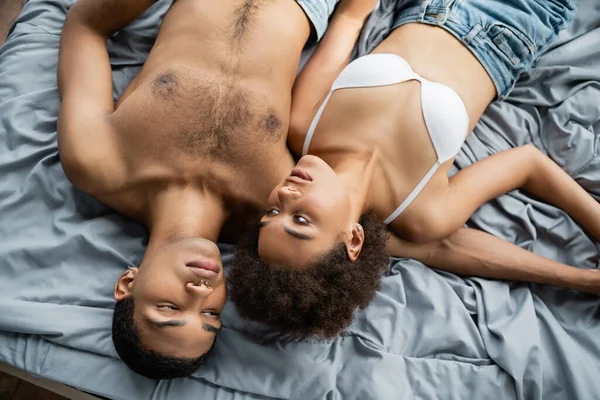 Vue du dessus de la femme afro-américaine en soutien-gorge blanc couché près de l'homme musclé torse nu sur le lit — Photo de stock