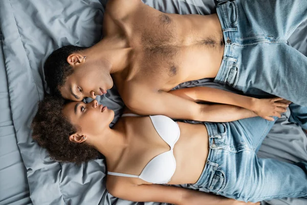 Top view of shirtless african american man and woman in bra lying face to face and holding hands — Stock Photo