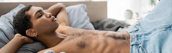 Hombre afroamericano sin camisa con piercing durmiendo en la cama en casa, pancarta - foto de stock