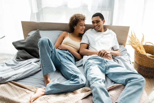 Young african american couple in jeans smiling while lying on bed — Stock Photo