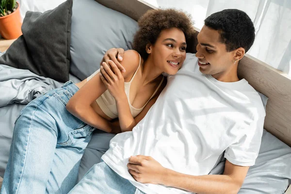 Высокий угол обзора молодой африканской американской пары, улыбающейся друг другу и обнимающейся на кровати — стоковое фото