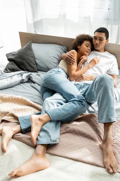 Longitud completa de la pareja afroamericana descalza en jeans abrazándose mientras está acostado en la cama con los ojos cerrados - foto de stock