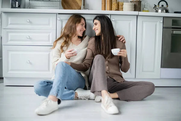 Vista lateral de mujeres lesbianas sosteniendo tazas de café mientras están sentadas en el suelo en la cocina - foto de stock