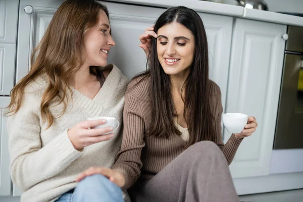 Молодая женщина держит чашку кофе и регулирует волосы партнера на кухне — стоковое фото
