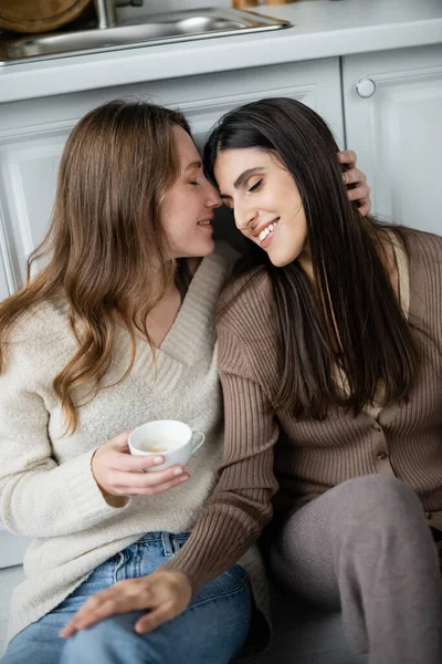 Lesbian woman in sweater hugging partner with cup of coffee in kitchen — Stock Photo