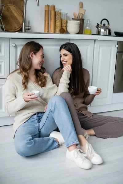 Молоді лесбійки тримають чашки кави, розмовляючи на підлозі на кухні — стокове фото