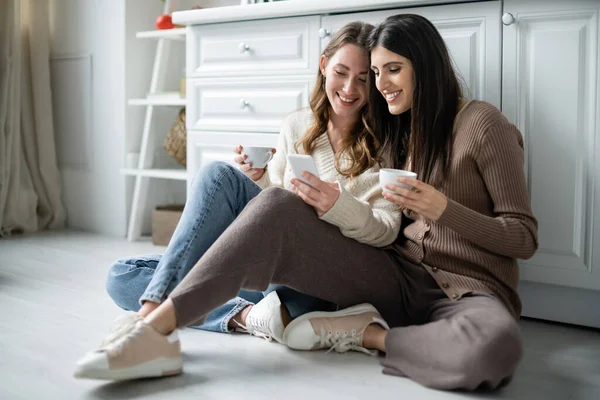 Lesbian women holding cups of coffee and using smartphone on floor in kitchen — Stock Photo