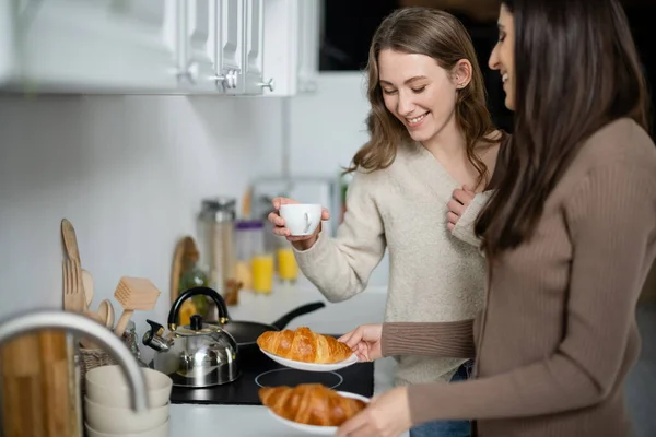 Улыбающаяся женщина в свитере держит чашку кофе рядом с подругой с круассанами на кухне — стоковое фото